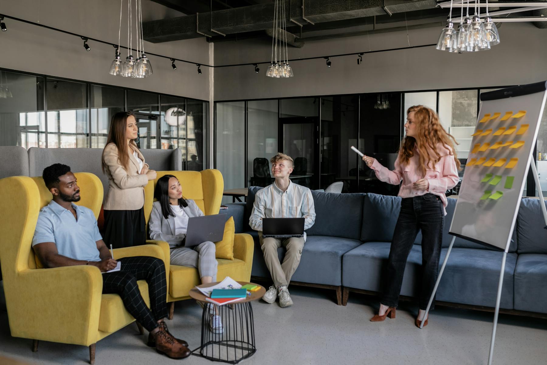 Marketing team in strategy planning session with woman presenting at whiteboard covered in sticky notes while colleagues listen. Image by A Darmel (https://www.pexels.com/@a-darmel)